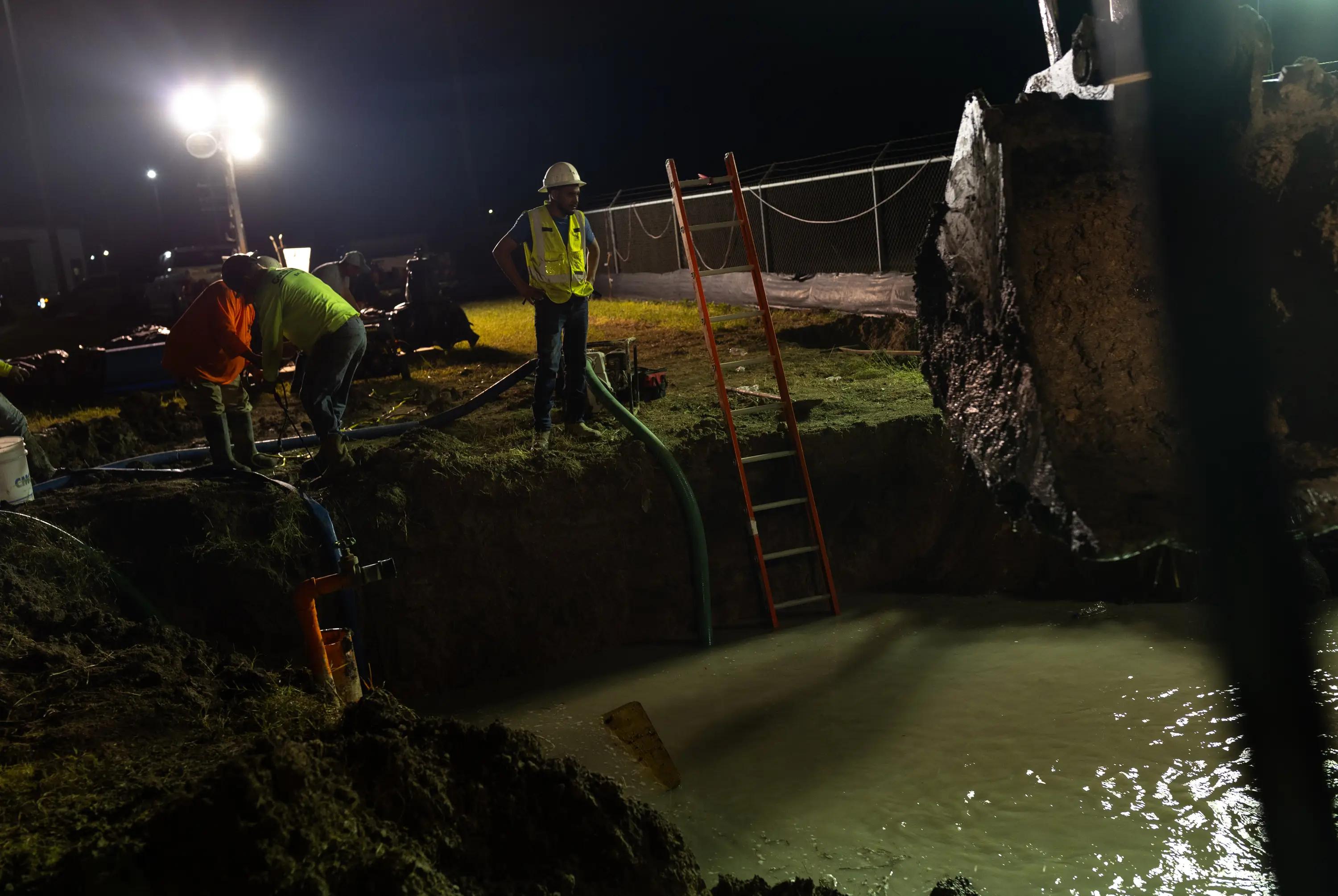 A construction workers stands next to a pit filled with water, illuminated by a light.