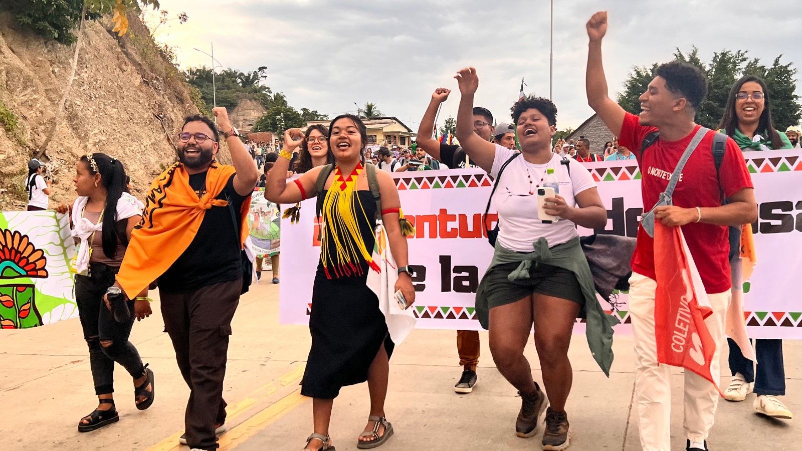 A group of young people holding signs in Spanish with their arms raised walk down a dusty road.