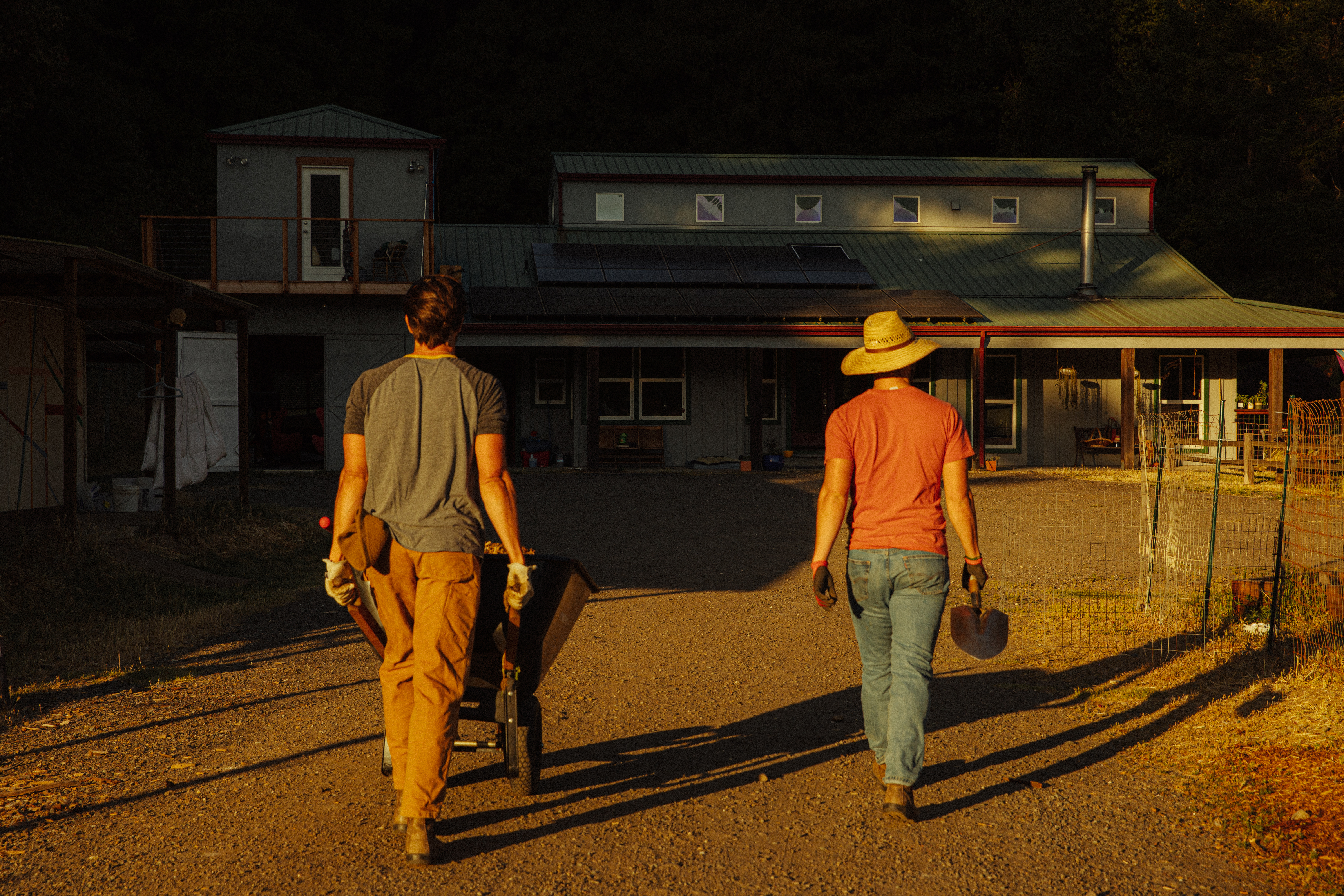 Two men walk toward a farmhouse with solar panels on the roof. The man on the left is pushing a wheelbarrow while the one on the right carries a shovel.