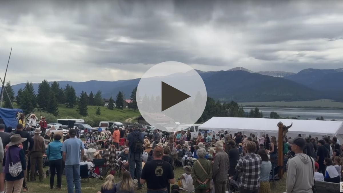 A still image with a play button overtop of it shows a large group of people assembled outside with green trees and tall mountains in the distance