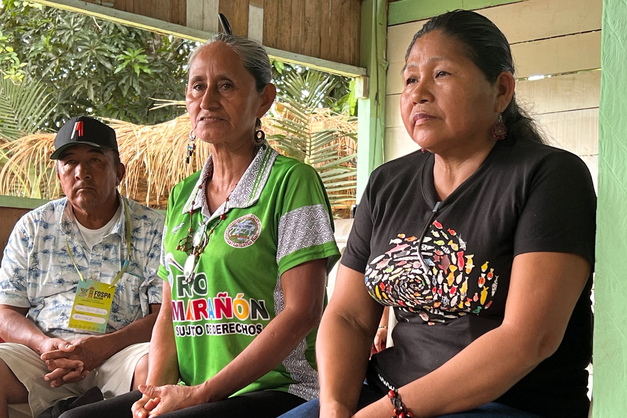 A man sits next to two women on a porch surrounded by tropical plants in the background.