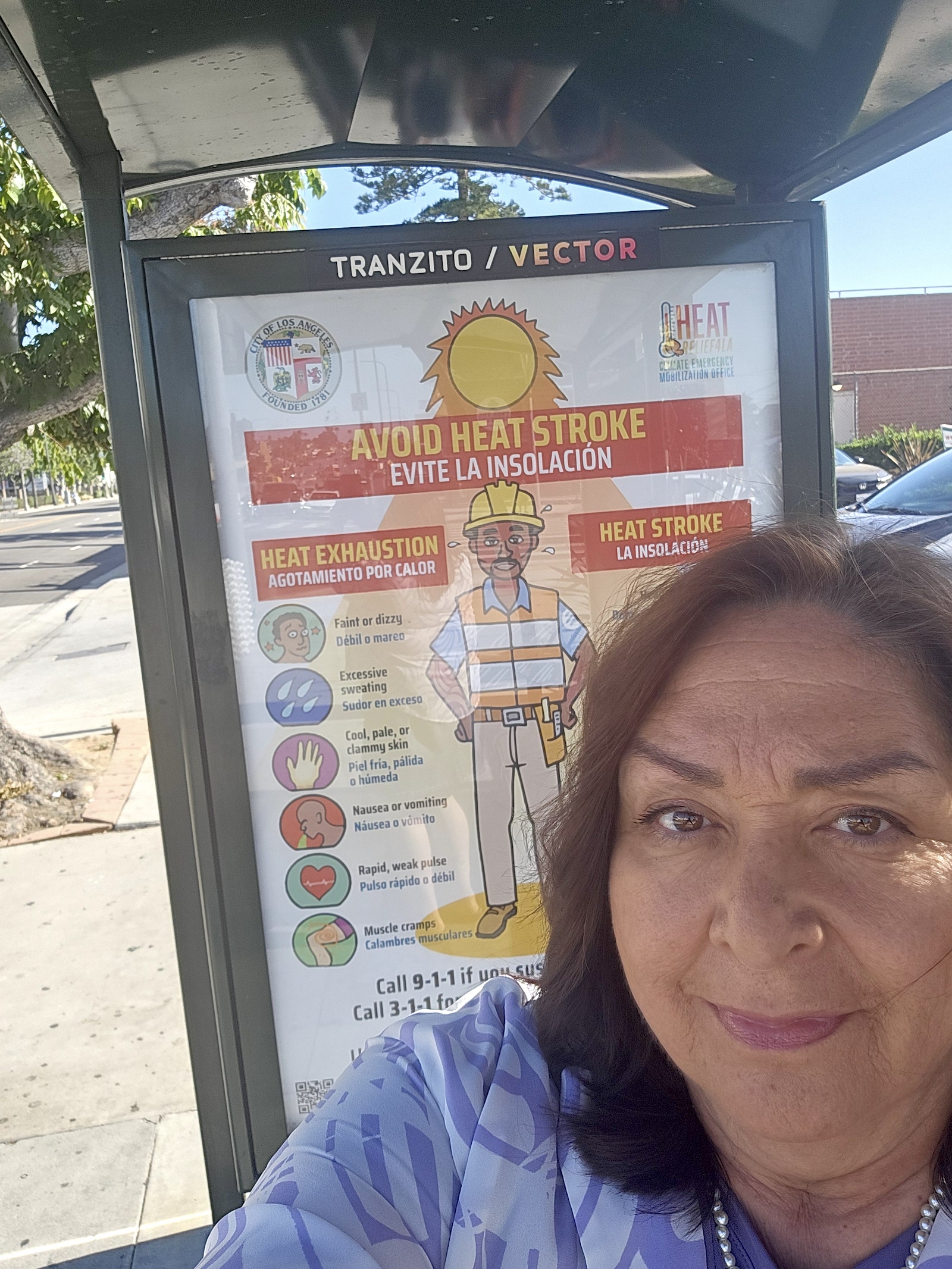 A woman stands selfie-style in front of a red and white bus stop poster with the header 'avoid heat stroke'